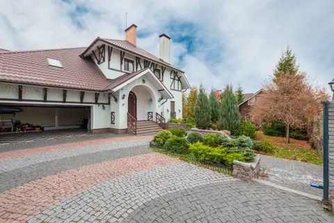 A quaint suburban house with a beautiful garden and brick driveway under a partly cloudy sky.