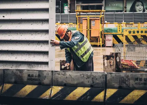 A construction worker in a reflective vest and hard hat at a busy urban construction site.