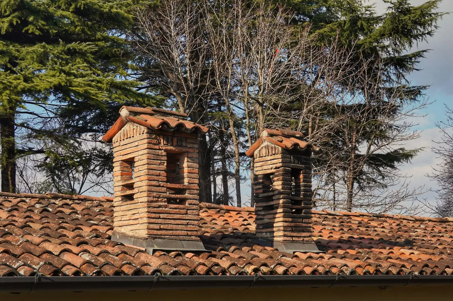 Two rustic brick chimneys on a red-tiled roof with a winter forest backdrop.
