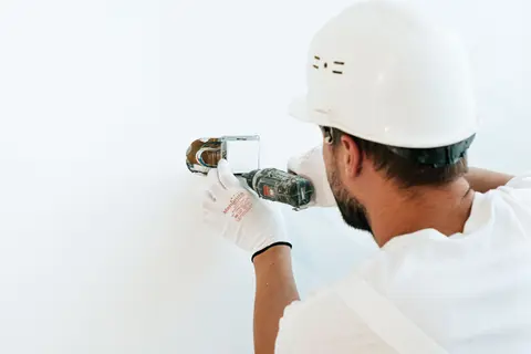 Construction worker wearing helmet and gloves uses a power tool to fix a wall.