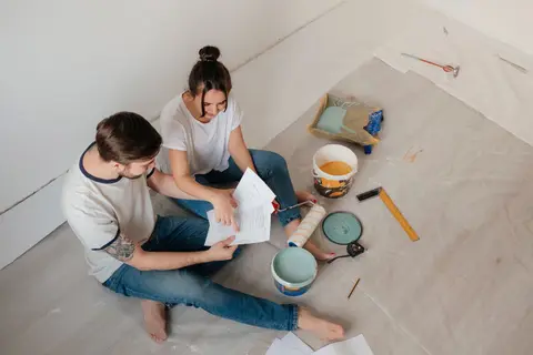 A couple sitting on the floor, reviewing documents amidst home renovation materials.