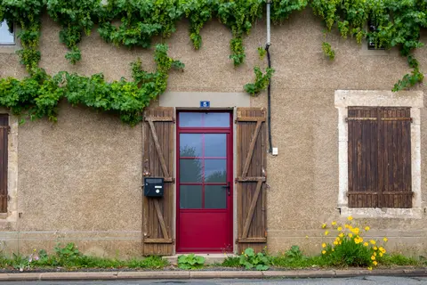 Rustic house exterior featuring a vibrant red door, wooden shutters, and ivy-clad walls.