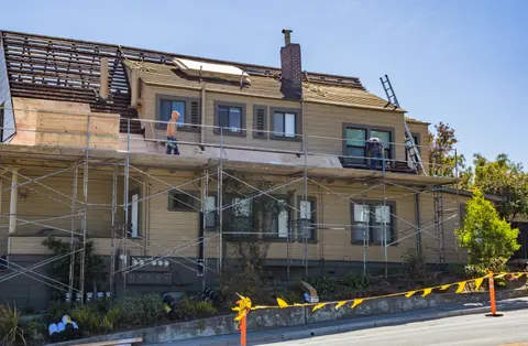 A house under renovation with scaffolding up and roofers working on the roof covering.