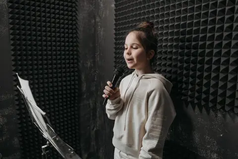 A young girl sings into a microphone in a soundproof room with acoustic panels around her.